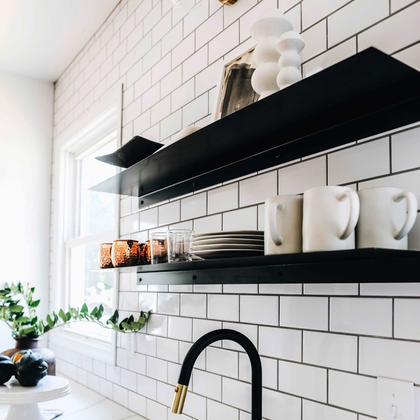 two black floating metal wall shelves mounted on white kitchen tile backsplash there are mugs, plates, and decor