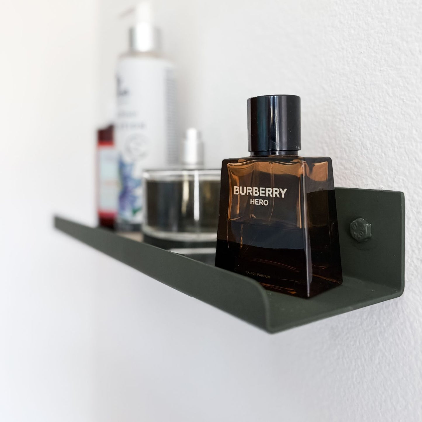 A small olive green metal shelf ledge mounted on a white wall with lotion bottles and cologne being displayed