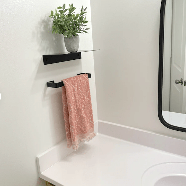 White bathroom with a black hand towel bar mounted on wall next to sink with a small floating metal shelf above with a plant.