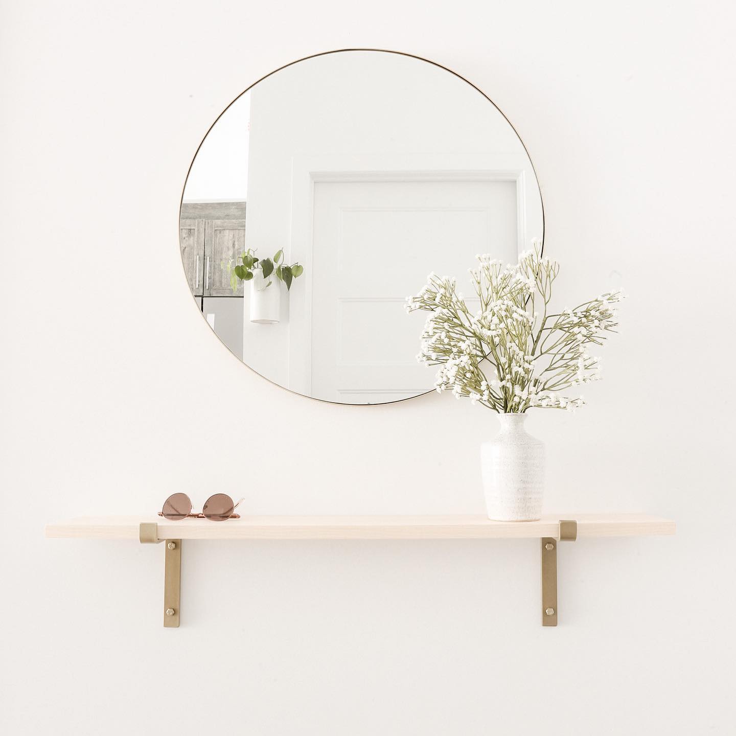 Entryway small wood shelf on white wall mounted with brass z style brackets. There is a round gold mirror above.