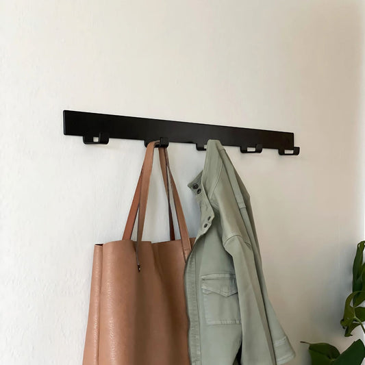 black metal coat rack with four hooks mounted on white wall with jacket and brown bag hanging