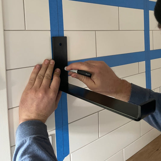 man holding black metal J bracket up to kitchen wall and marking hole placement on white tike backsplash for installation