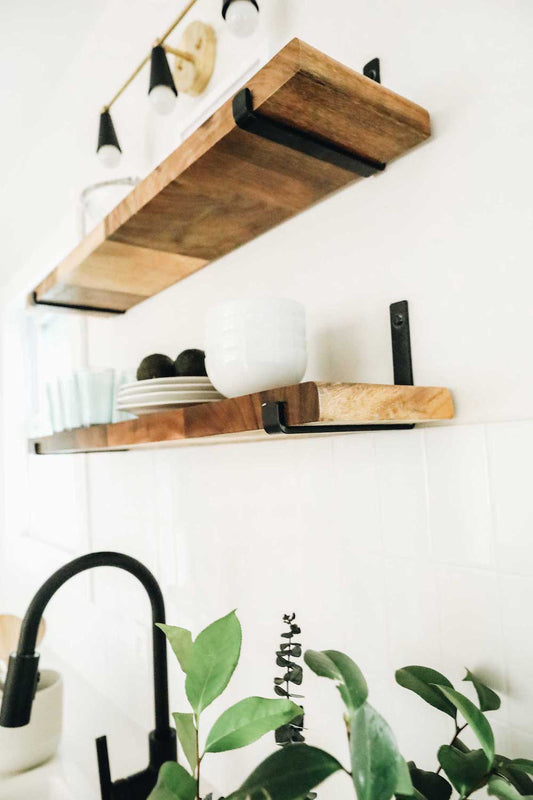Kitchen with two wood shelves mounted on white  wall with black J style hook shelf brackets. There is a faucet and plant