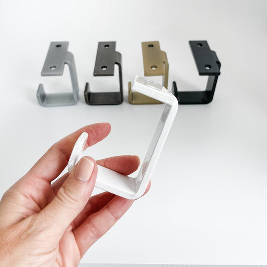 woman's hand holding white under mount J shaped hook there are more hooks in the background in silver, steel, brass, black