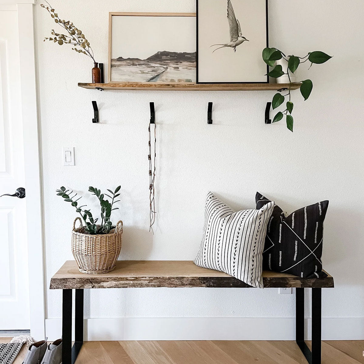 entryway with four black metal hooks mounted on wall underneath wood shelf. There is also a wood bench with pillows and plant