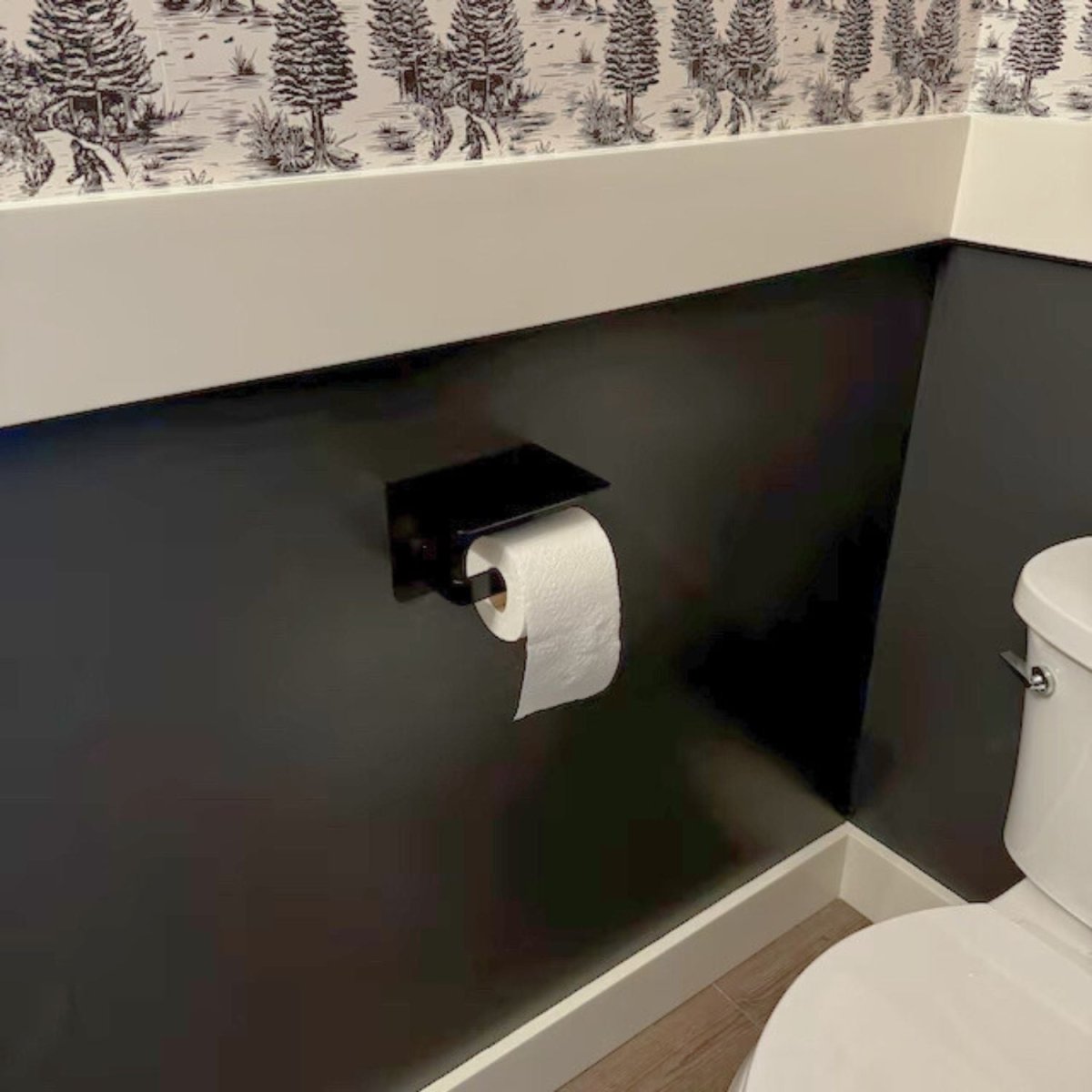 black bathroom with wall mounted metal toilet paper holder with attached shelf. There is a white trim board and black and white wallpaper above. 
