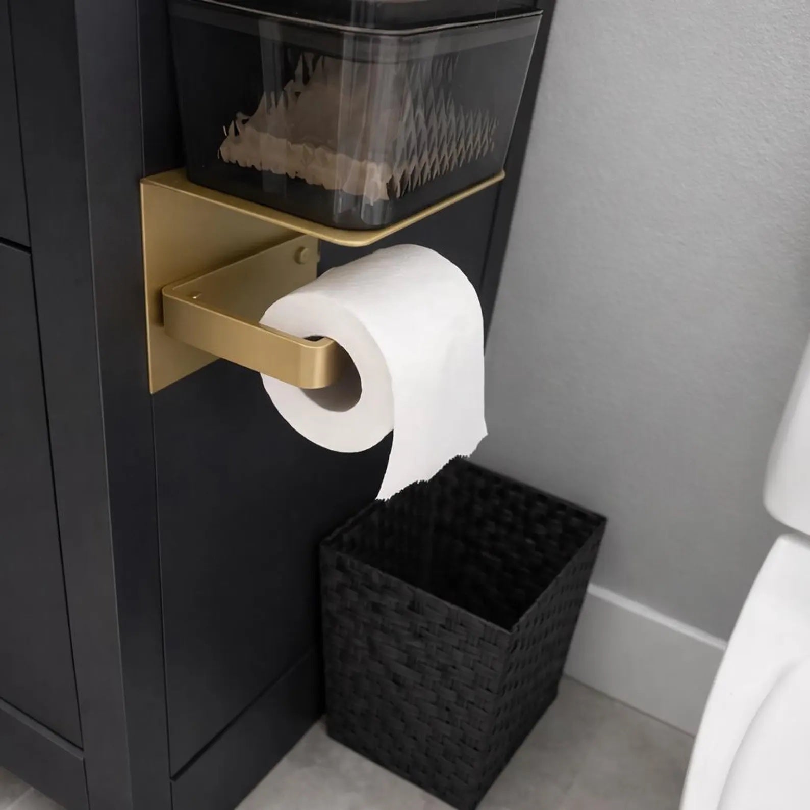 Black bathroom cabinets with a brass toilet paper holder and a shelf with a large tissue roll mounted on the side