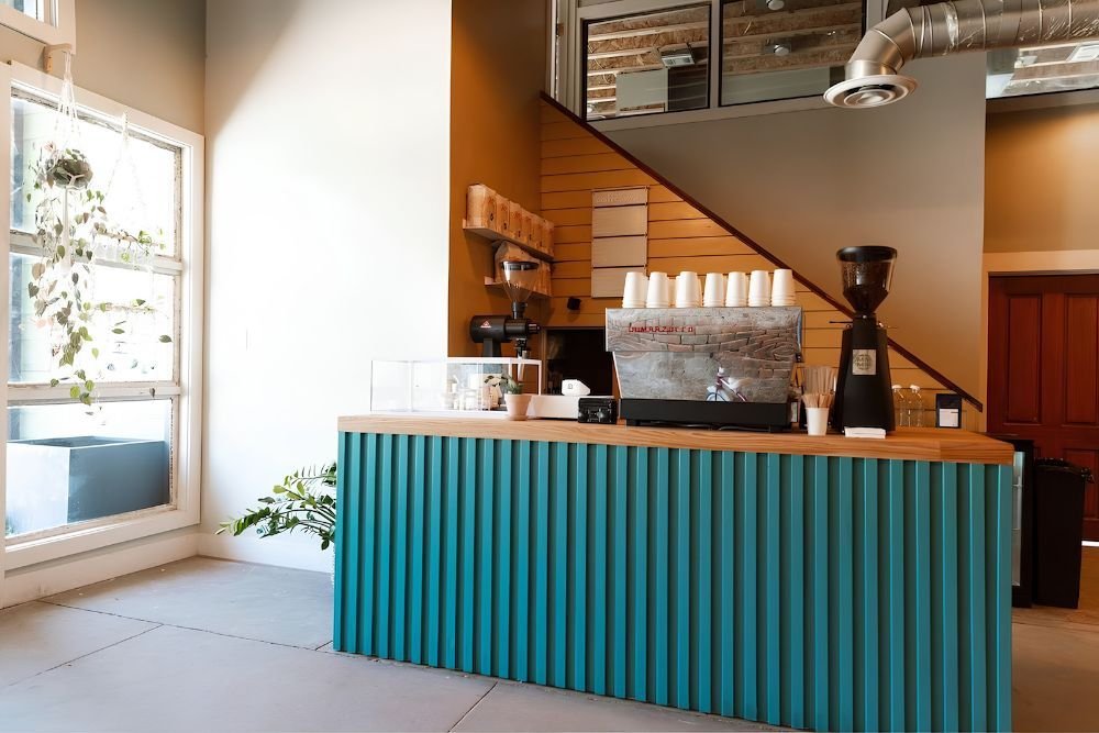 Modern coffee shop interior with a teal counter and coffee-making equipment. There are floating display shelves on wall.