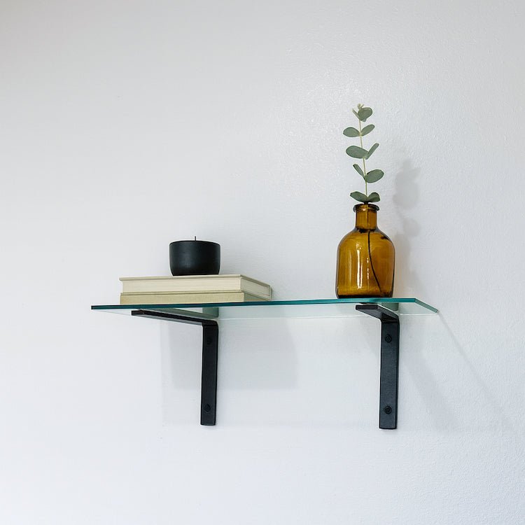 Glass shelf with black brackets holding a vase, books, and a candle against a white wall.