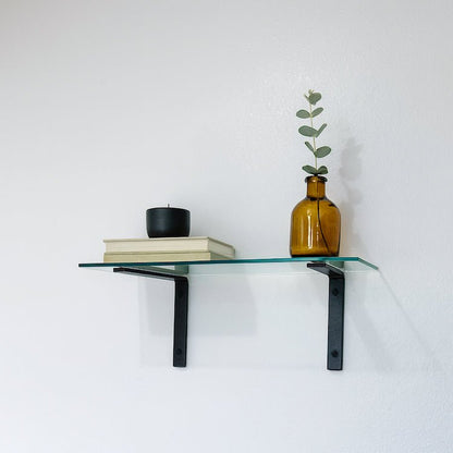 Glass shelf with black brackets holding a vase, books, and a candle against a white wall.