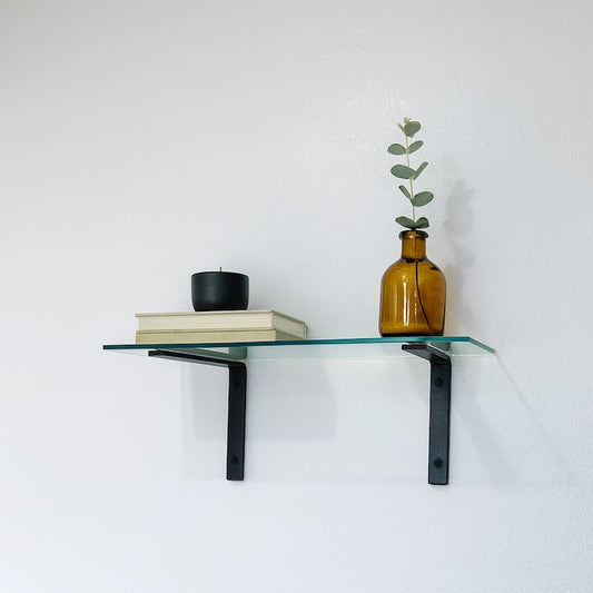 Glass shelf with black brackets holding a vase, books, and a candle against a white wall.
