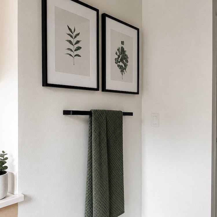 Green towel hanging on a black rack with framed botanical prints on a white wall.