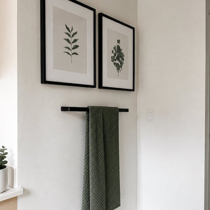 Green towel hanging on a black rack with framed botanical prints on a white wall.