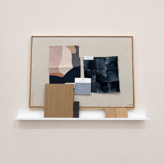 An abstract framed wall art piece displayed on a white shelf ledge against a warm beige wall. The artwork features a beige linen-like background with geometric and organic shapes in navy, blush pink, tan, and charcoal, framed in a thin light wood border.