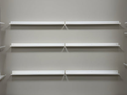 Six wall mounted white metal display ledge shelves on gray wall. Each shelf rail is empty.